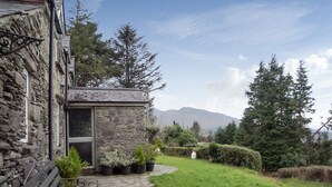 A stone building with a small entrance, a stone pathway leading to a bench, and a grassy area with trees and mountains in the background. - Angelas Farmhouse (Lauragh)