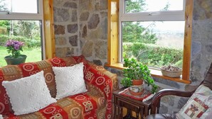 A cozy living room with a stone wall, a patterned sofa, a wooden chair, and a view of greenery through large windows. - Angelas Farmhouse (Lauragh)