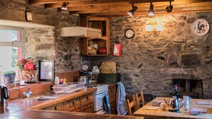 A rustic kitchen with stone walls, wooden beams, a stove, and a dining area with a table and chairs. - Angelas Farmhouse (Lauragh)