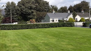 A residential house with a well-maintained lawn, a white fence, and a large tree in the background. - Garden Cottage (Clogheen)