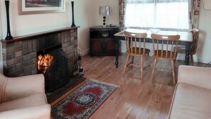 A living room with a fireplace, a dining table with chairs, a window with curtains, and a framed picture on the wall. - Garden Cottage (Clogheen)