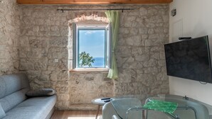 Ein Zimmer mit Steinwänden und Holzboden, ein graues Sofa, ein kleiner Tisch mit Fernbedienung, eine Glasdusche und ein an der Wand befestigter Fernseher.