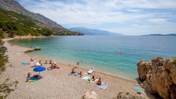 Ein Strand mit badenden Personen, blauem Himmel und Bergen im Hintergrund.