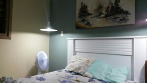 A bedroom with a bed, a standing fan, and a painting on the wall. - Cozy in Floripa (Florianópolis)