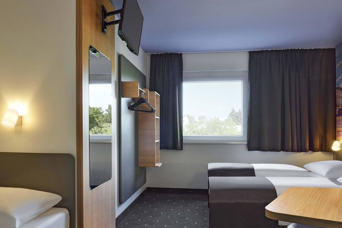 Quarto de hotel com cama, sofá, mesa pequena, televisão na parede e janela com cortinas.