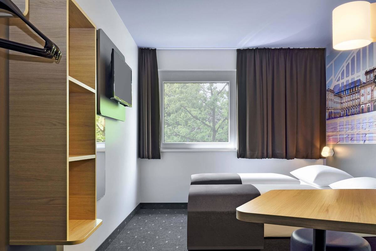 Quarto de hotel com cama, escrivaninha, cadeira, televisão e janela com cortinas.