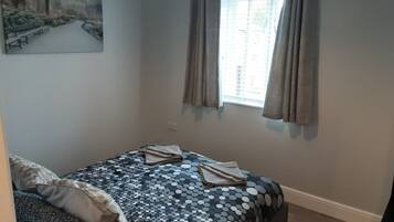 A bedroom with a bed, pillows, a window with curtains, a painting on the wall, and a carpeted floor.