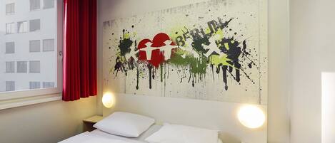 Une chambre d’hôtel avec deux lits, un rideau rouge, un mur orné d’une œuvre d’art abstraite, et une fenêtre donnant sur des immeubles.