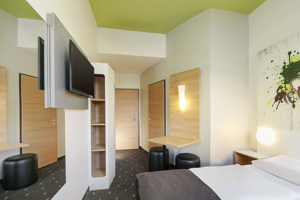 Ein modernes Hotelzimmer mit einem Bett, einem Flachbildfernseher an der Wand, einem kleinen Schreibtisch und einer Wandleuchte.