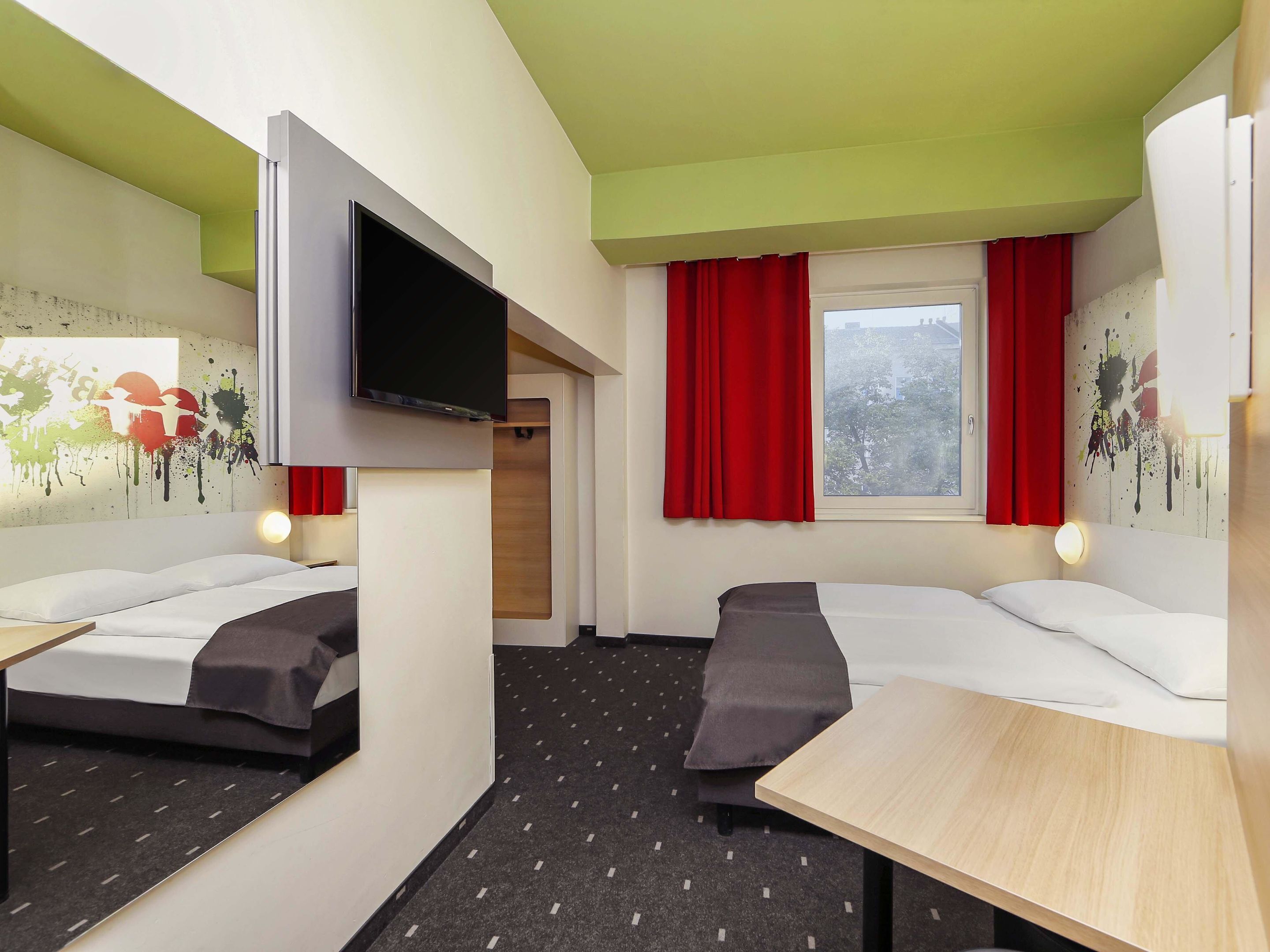 Habitación de hotel con dos camas, televisor, mesa y ventana con cortinas rojas.