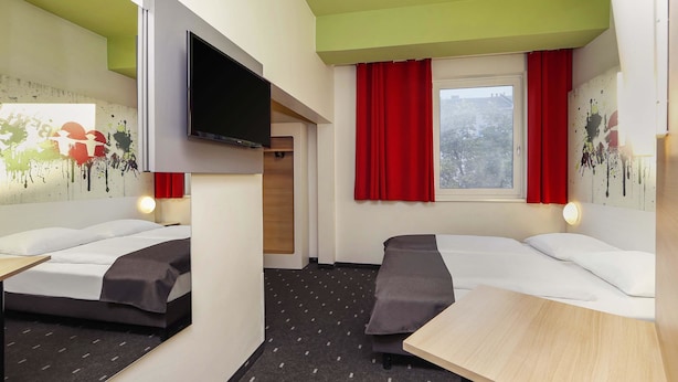 A hotel room with two beds, a TV, a table, and a window with red curtains.