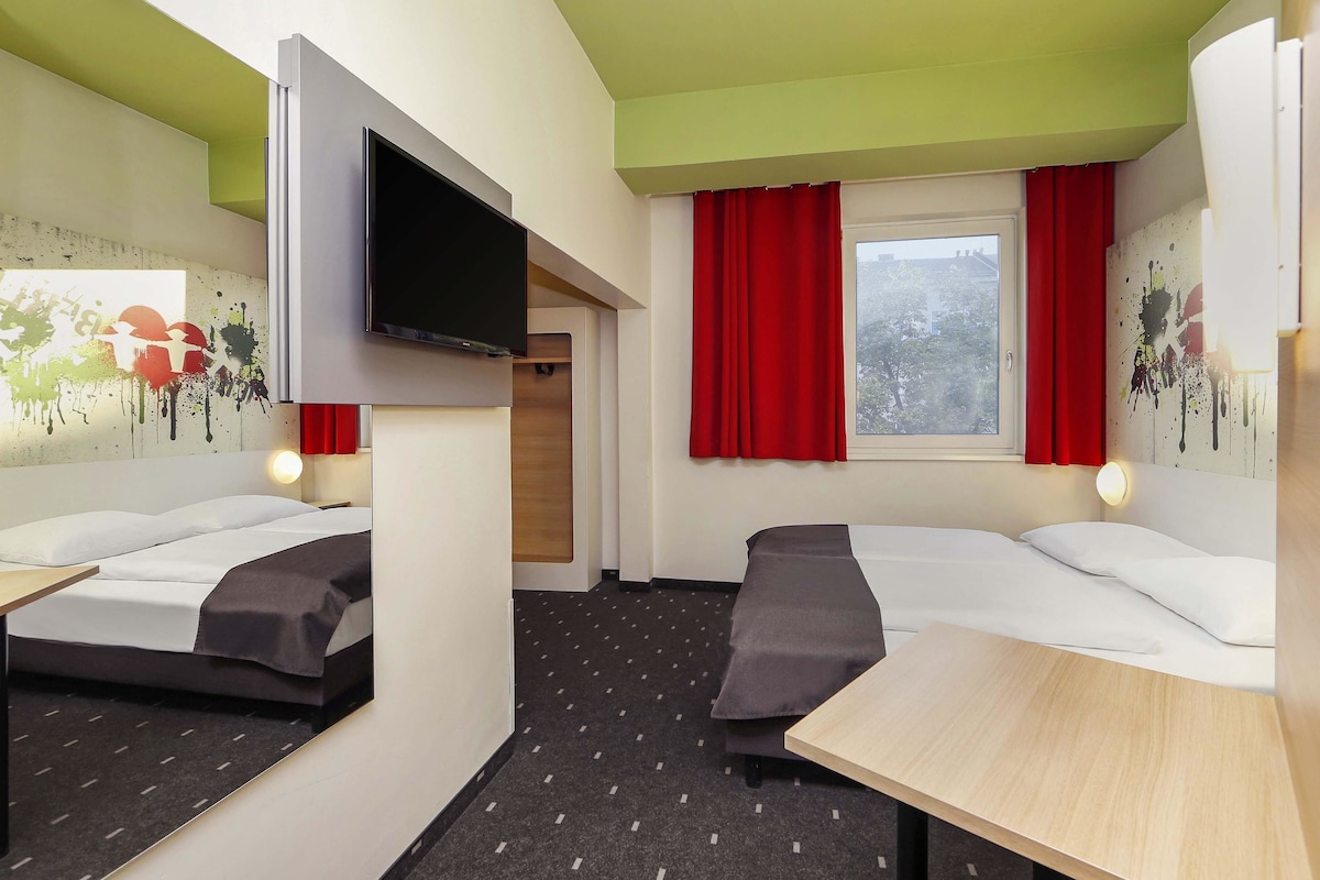 Ein Hotelzimmer mit zwei Betten, einem Fernseher, einem Tisch und einem Fenster mit roten Vorhängen.