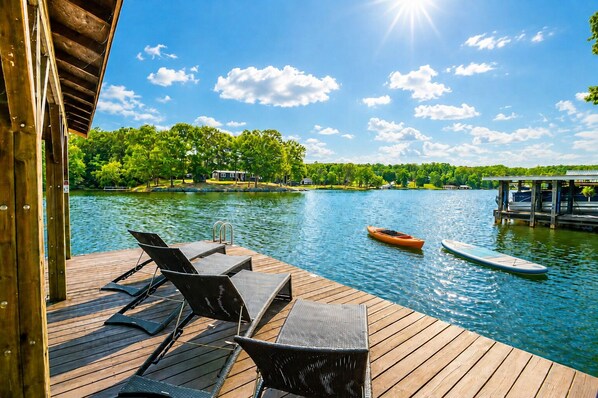 A wooden deck with lounge chairs and a kayak on a calm lake, surrounded by greenery and a clear sky.