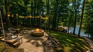 A fire pit surrounded by chairs and a string of lights, set against a backdrop of trees and a body of water. - Lake-Front - Hot Tub, Large Dock, Fire Pit, Deck (Roxboro)