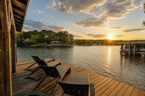 Een houten terras met lounge fauteuils en een tafel, met uitzicht op een meer bij zonsondergang.