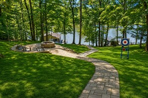 Un espace extérieur pavé, aménagé avec un foyer, des sièges et une cible, situé près d’une étendue d’eau.