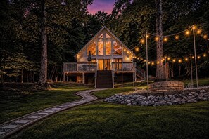 A cabin with a deck, surrounded by trees and a stone wall, with a pathway leading to it.