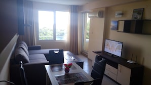A living room with a sofa, a dining table with a vase of flowers, a TV on a stand, and a balcony with curtains. - Nikea Park (Varna)