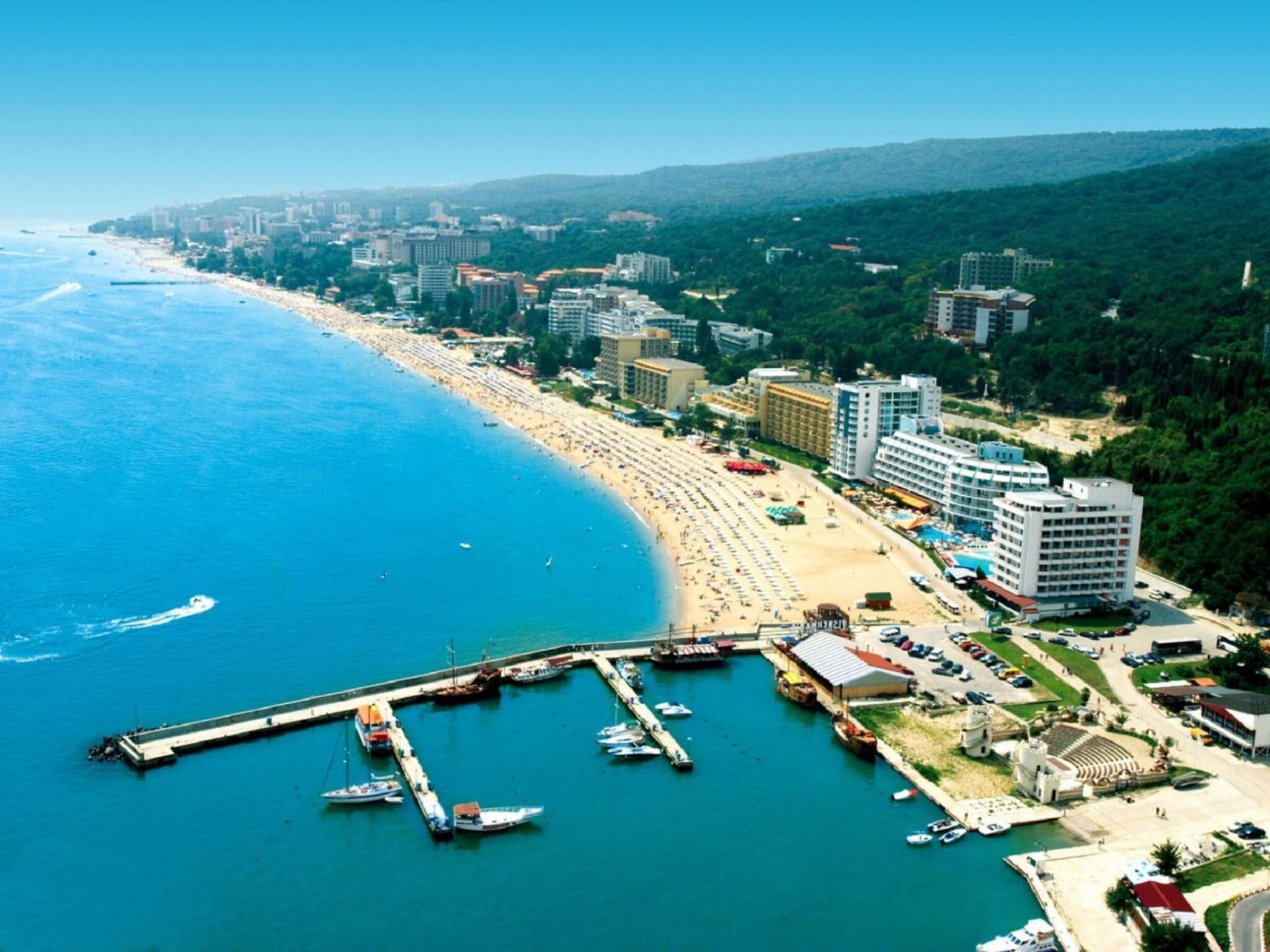 An aerial view of a coastal city with a long beach, a marina with boats, and several high-rise buildings.