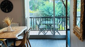 Un coin repas avec une table et des chaises en bois, une horloge murale et une vue sur un balcon aménagé avec du mobilier d’extérieur.