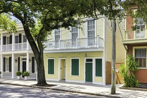 A row of two-story houses with balconies, a tree in the foreground, and a sidewalk.
