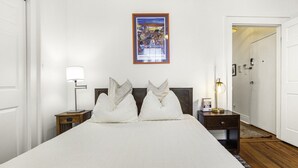Habitación de hotel con una cama grande, dos mesitas de noche, una mesita auxiliar, un cuadro en la pared y una puerta que conduce a otra habitación.