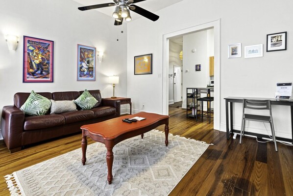 A living room with a brown leather sofa, a wooden coffee table, and a ceiling fan. There are framed pictures on the walls and a dining area in the background.