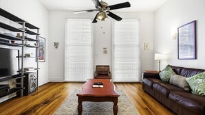 Una sala de estar con piso de madera, un sofá de cuero marrón, una mesa de centro, un mueble para televisor y un ventilador de techo.