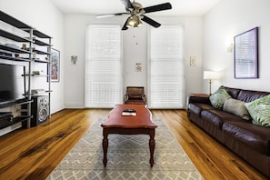 A living room with a wooden floor, a brown leather sofa, a coffee table, a TV stand, and a ceiling fan.