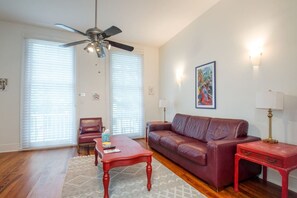 Sala de estar com sofá de couro vermelho, mesa de centro de madeira, mesa lateral vermelha, ventilador de teto e um quadro na parede.