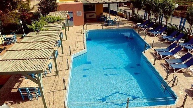 En rektangulär swimmingpool med ett kaklat poolområde, omgiven av solstolar och en möblerad, överbyggd sittplats.