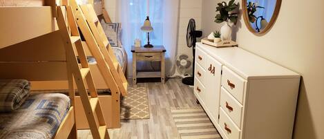 Ein Schlafzimmer mit Etagenbett, Nachttisch mit Lampe, Kommode, Spiegel und einem Fenster mit Vorhängen.