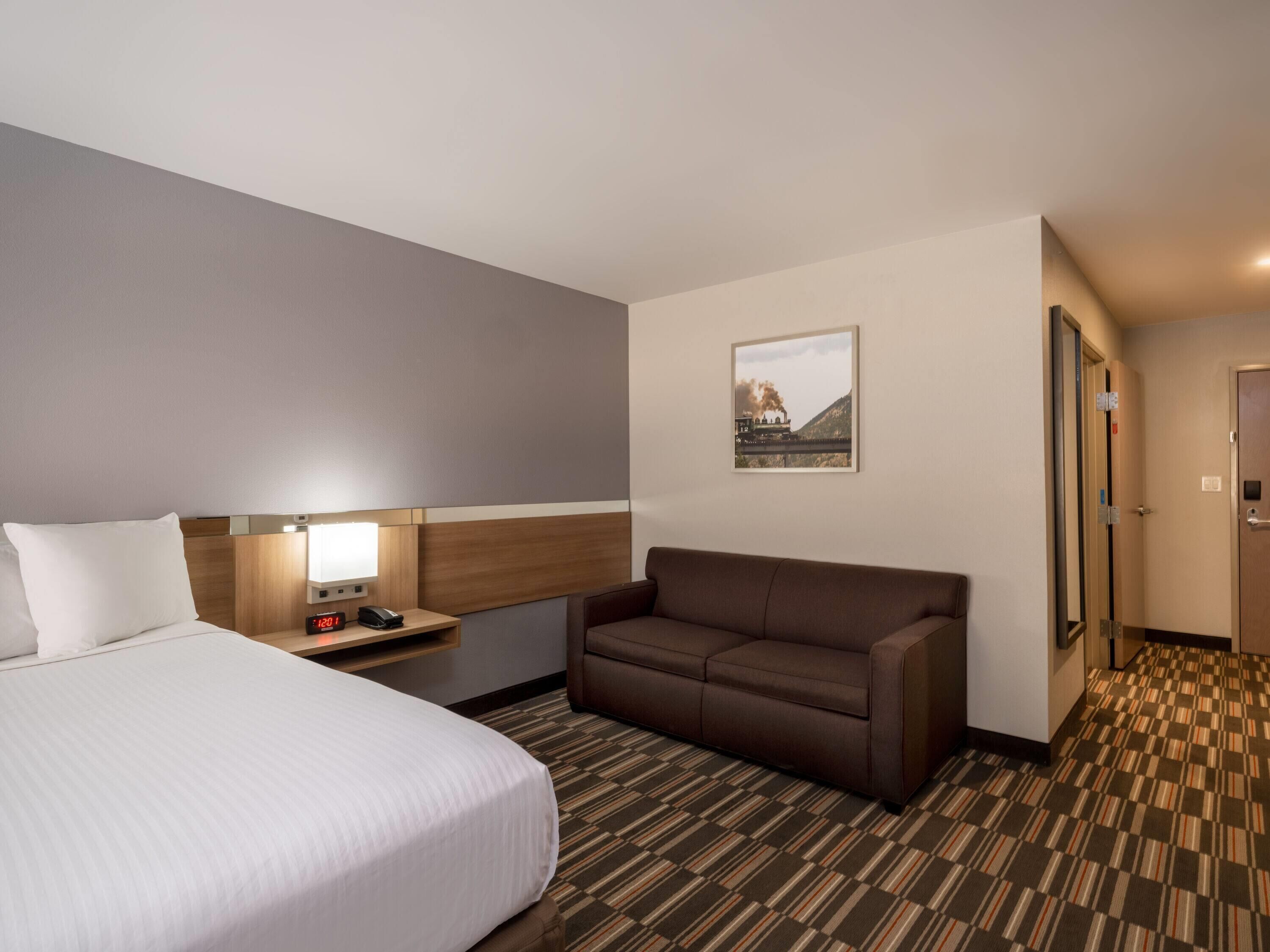 A hotel room with a bed, a nightstand with a lamp, a brown sofa, a framed picture on the wall, and a patterned carpet.