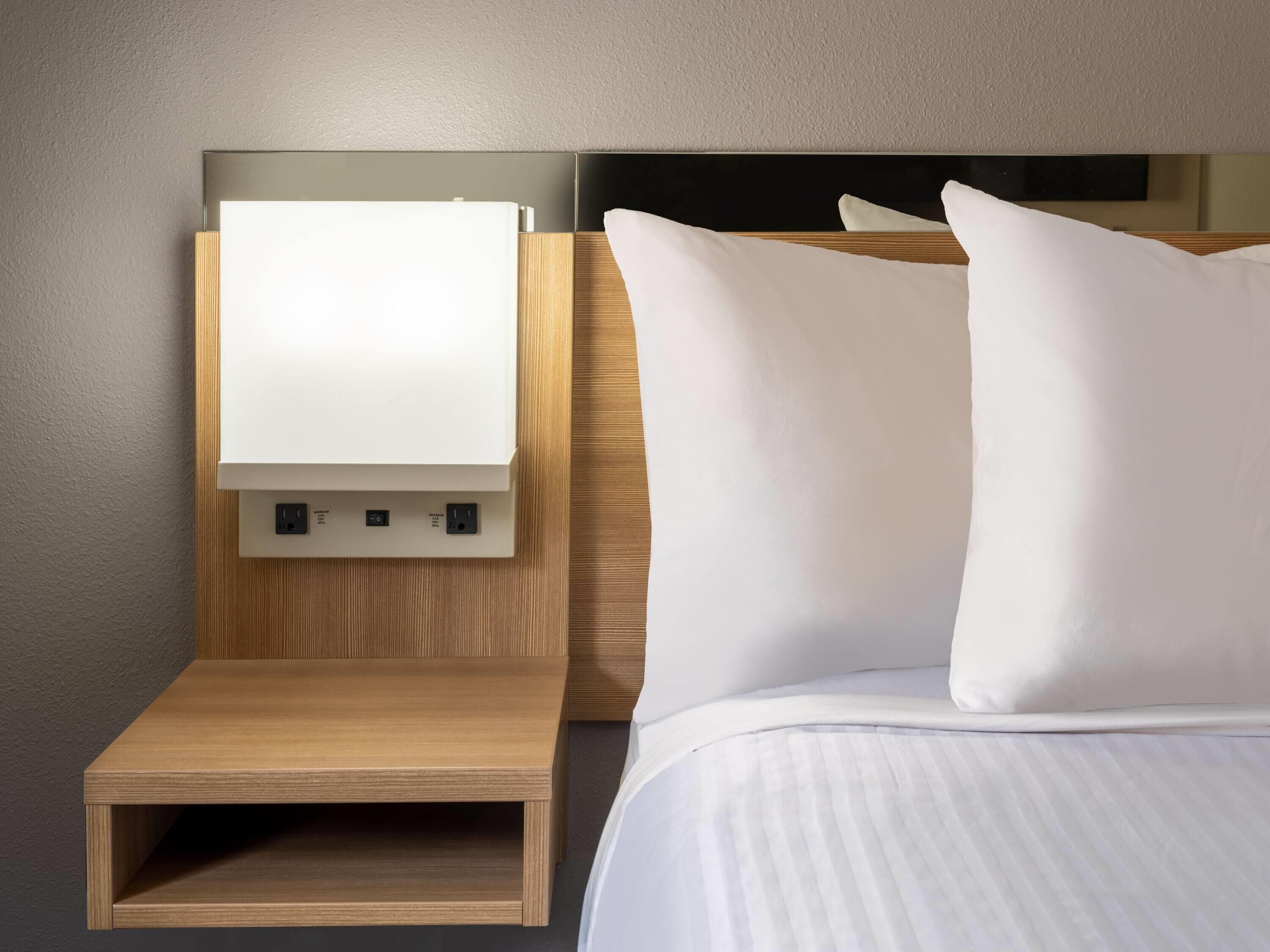 A neatly made bed with white linens and pillows, a wooden bedside table with a flat-screen TV mounted above it, and a power outlet on the wall.