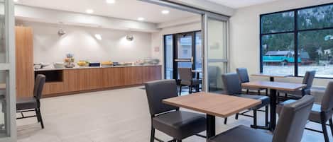A modern dining area with tables and chairs, a counter with food items, and large windows offering a view of the outdoors.