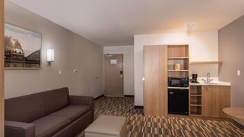 A hotel room with a brown sofa, a small ottoman, a kitchen area with a microwave, a sink, and a cabinet.