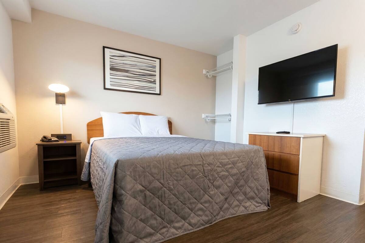 Una habitación de hotel con cama, mesita de noche, lámpara, televisor y una obra de arte colgada en la pared.