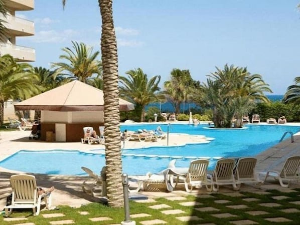 A pool area with lounge chairs and a small building, surrounded by palm trees. - Riviera Beach Apartments (Pilar de la Horadada)