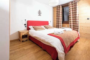 Une chambre avec un plancher en bois, une couvre-lit rouge, deux oreillers, une table de chevet et une fenêtre avec des rideaux.