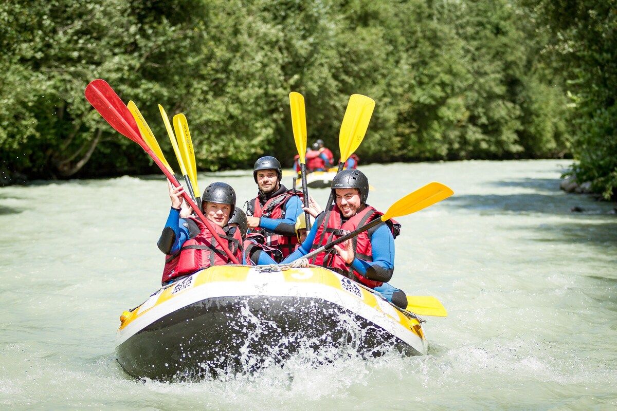 Un groupe de personnes se trouve dans un radeau, pagayant avec des pagaies, portant des casques et des gilets de sauvetage, sur une rivière bordée de verdure.
