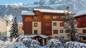 Una baita di legno con ampie finestre e un tetto imbiancato dalla neve, circondata da alberi carichi di neve e montagne sullo sfondo.