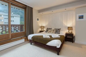 A hotel room with a large bed, two bedside tables, a window with a view of buildings, and a radiator. - Chamois Apartment, amazing views Chamonix All Year (Chamonix)
