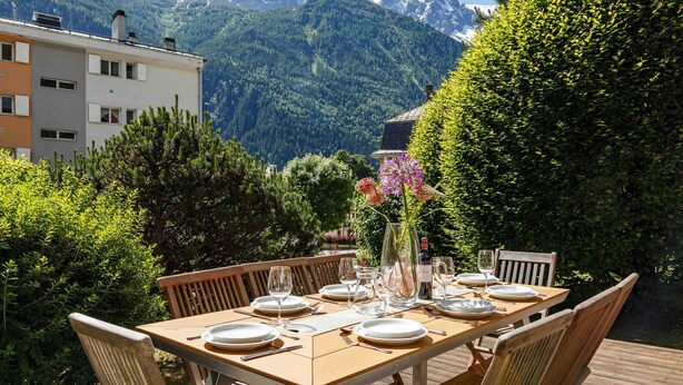 Area pranzo all'aperto con tavolo imbandito per quattro persone, circondato da sedie di legno e vista sulle montagne.