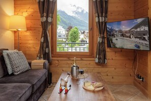 A cozy room with a wooden interior, a sofa, a table with a bottle, glasses, a wooden horse sculpture, and a TV displaying a scenic view. - Les Sonnailles Appt, Skiing, Chamonix All Year (chamonix)