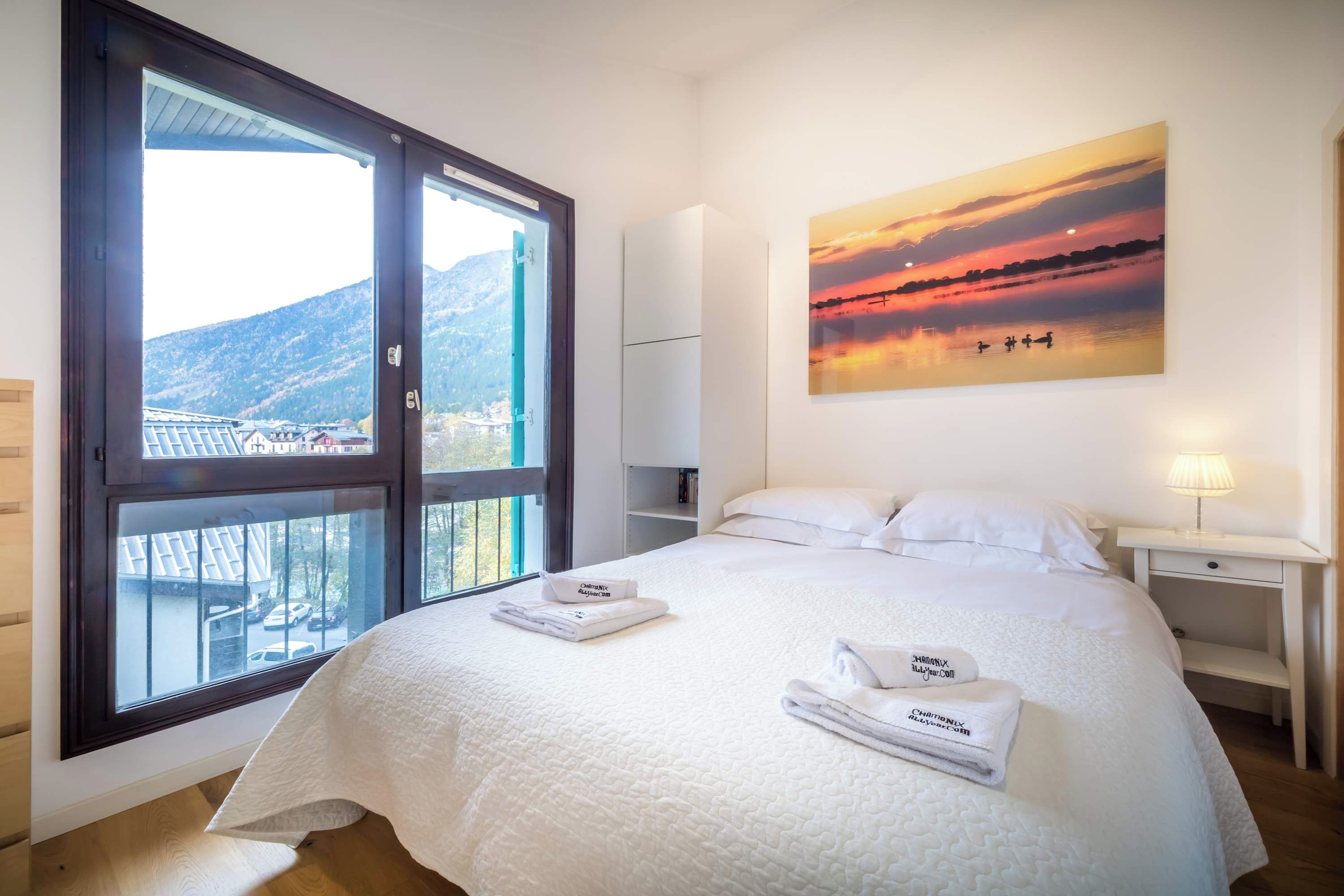 A hotel room with a bed, a nightstand, a painting of a sunset, and a view of a mountain through a large window.
