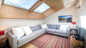 A modern living room with a grey sectional sofa, a patterned rug, wooden beams, and a mountain view through skylights. - Rupicapra apartment -  an apartment that sleeps 6 guests  in 3 bedrooms (Chamonix)