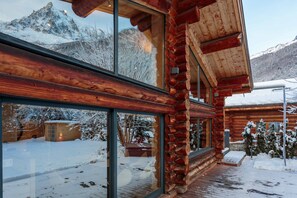 A log cabin with large windows, a wooden deck, and a snowy mountain backdrop.