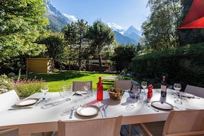 An outdoor dining area with a table set for a meal, surrounded by a lush garden and a mountain view.