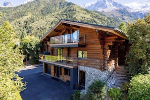 Une cabane en rondins dotée d’un balcon, avec des fondations en pierre et une vue sur les montagnes en toile de fond.
