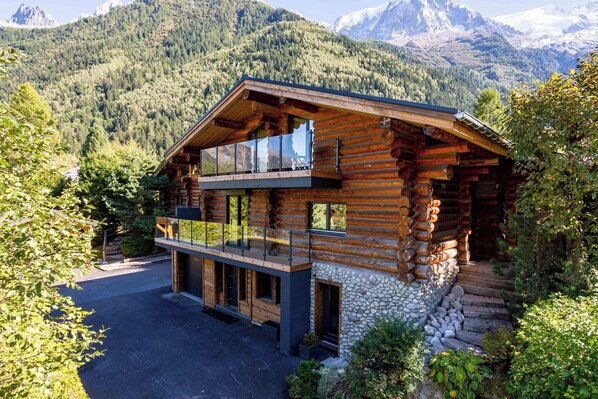 A log cabin with a balcony, stone foundation, and a mountain backdrop.